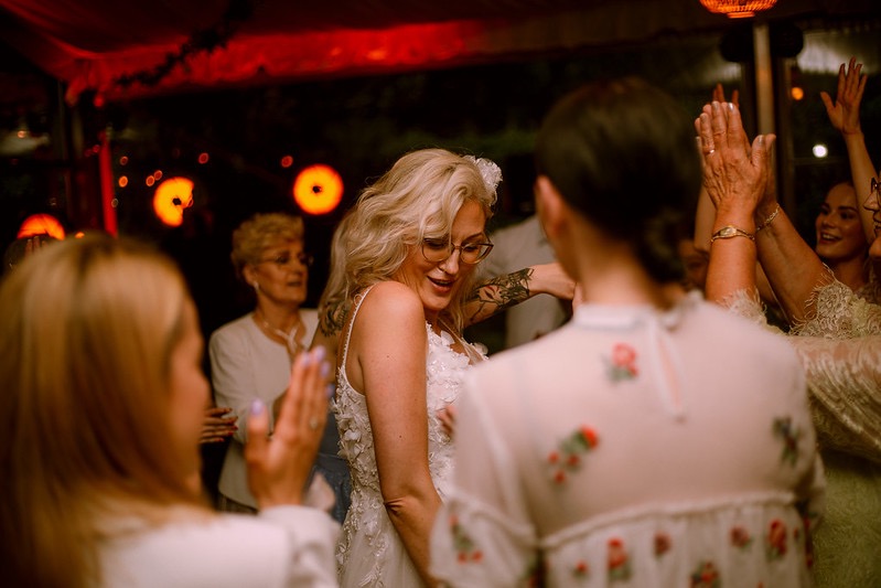 Uśmiechnięta panna młoda w okularach i sukni z aplikacjami kwiatowymi tańczy z gośćmi weselnymi w ciepłym, czerwonym oświetleniu.