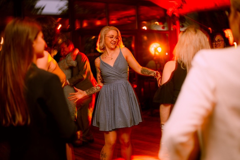 Kobieta w błyszczącej sukience tańczy na parkiecie podczas wydarzenia, otoczona innymi osobami, oświetlona czerwonym światłem.