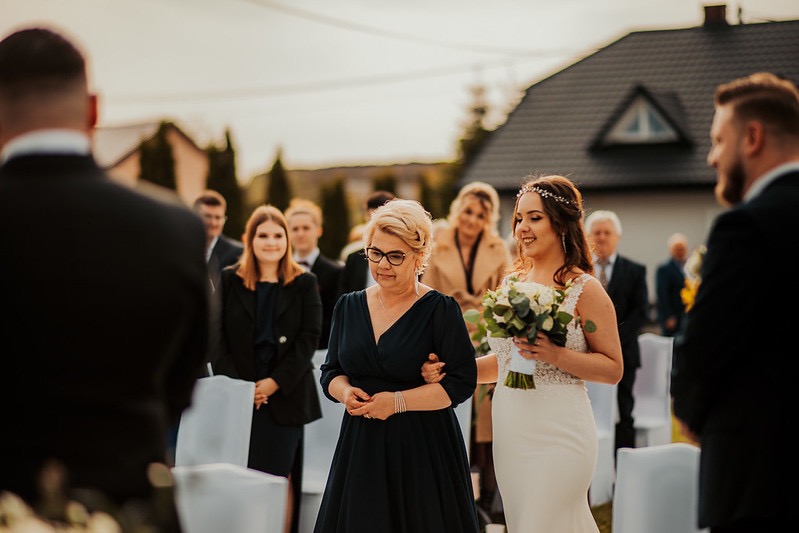 Panna młoda prowadzona do ołtarza przez matkę, w tle goście i dom z ciemnym dachem.