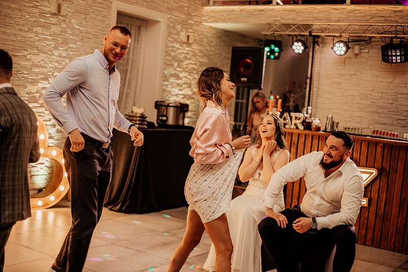 Rozbawiona panna młoda w białej sukni siedzi obok mężczyzny z brodą, podczas gdy goście weselni tańczą na parkiecie oświetlonym przez reflektory i dekoracyjne litery ze światłami