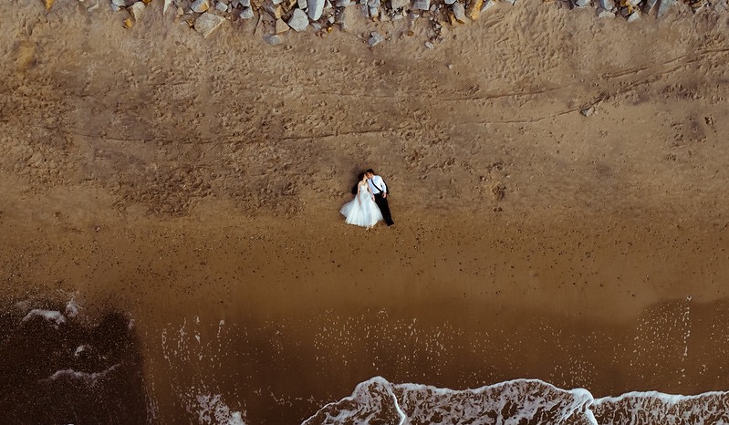 Para młoda leżąca na piaszczystej plaży, widok z góry ukazuje fale morskie obmywające brzeg i kamienisty klif w tle.