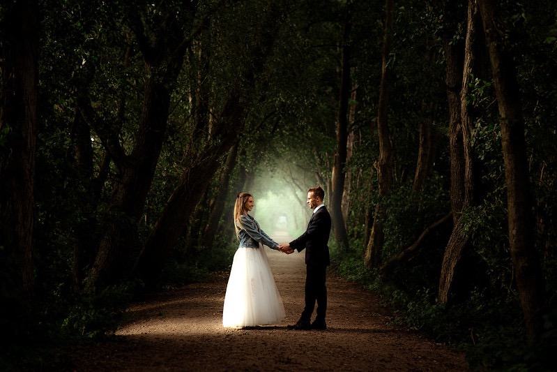 Para trzymająca się za ręce na leśnej ścieżce, panna młoda w białej sukni i jeansowej kurtce, pan młody w czarnym garniturze, mglista poświata w tle.