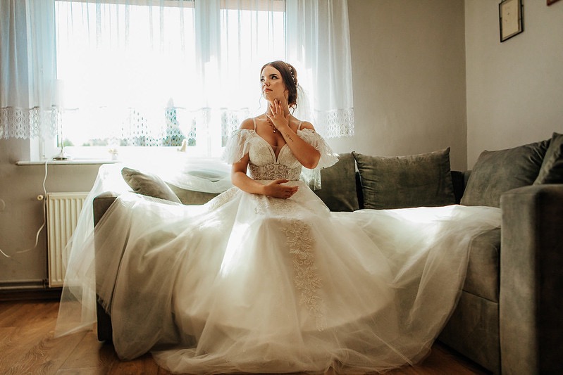 Panna młoda w białej sukni z koronkowymi zdobieniami siedzi na sofie, spoglądając w bok, z ręką na szyi, w tle okno z firankami.