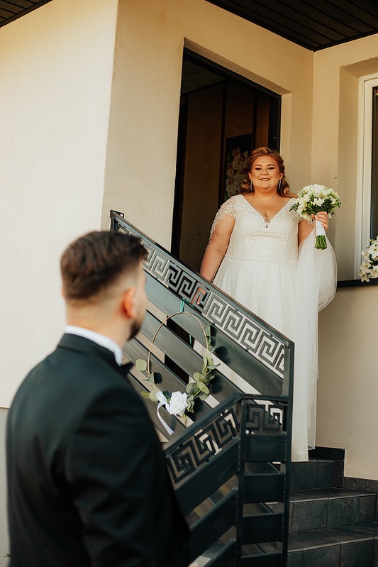 Panna młoda w koronkowej sukni ślubnej z bukietem białych kwiatów schodzi po schodach ozdobionych złotym kołem z kwiatami, widok zza pleców pana młodego w czarnym smokingu.