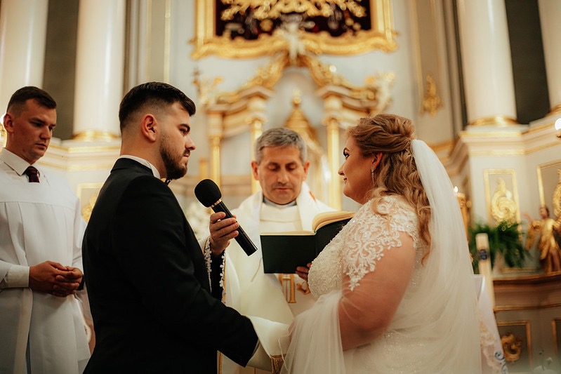 Para młoda podczas ceremonii ślubnej w kościele, ksiądz czyta z księgi, pan młody trzyma mikrofon.