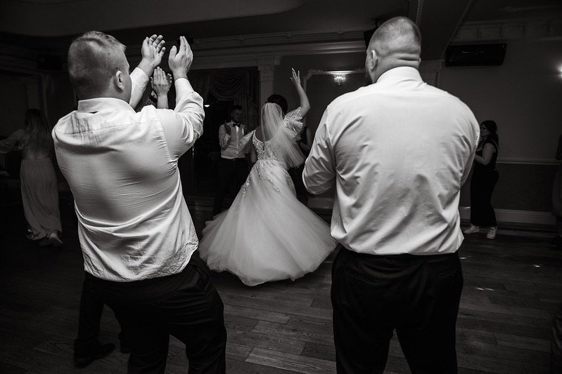 Czarno-białe zdjęcie tańczącej pary młodej w otoczeniu gości weselnych, z widocznymi rękami klaszczących osób w tle, parkiet z desek.