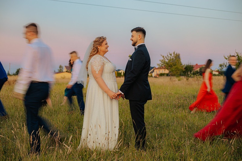 Para młoda trzymająca się za ręce na łące, w tle rozmyte postacie gości weselnych