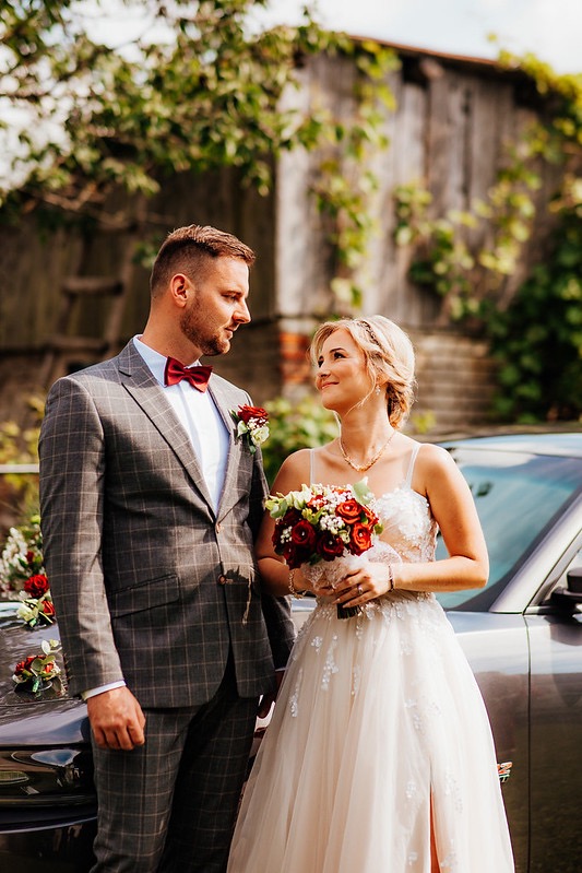 Para młoda w plenerze: Panna młoda w sukni z rozcięciem, trzyma bukiet czerwonych róż, patrzy na pana młodego w szarym garniturze w kratę i bordowej muszce, obok udekorowany samochód.