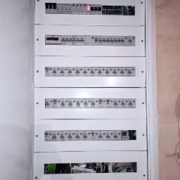 Rozdzielnica elektryczna w białej obudowie, widoczne zabezpieczenia nadprądowe, wyłączniki różnicowoprądowe i moduł pomiarowy z wyświetlaczem cyfrowym.