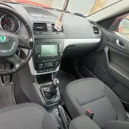 Wnętrze samochodu Skoda Yeti: deska rozdzielcza z nawiewami, radioodtwarzaczem i zawieszką zapachową, kierownica z logo marki, czarne fotele z czerwonymi pasami bezpieczeństwa, gumowe dywaniki.