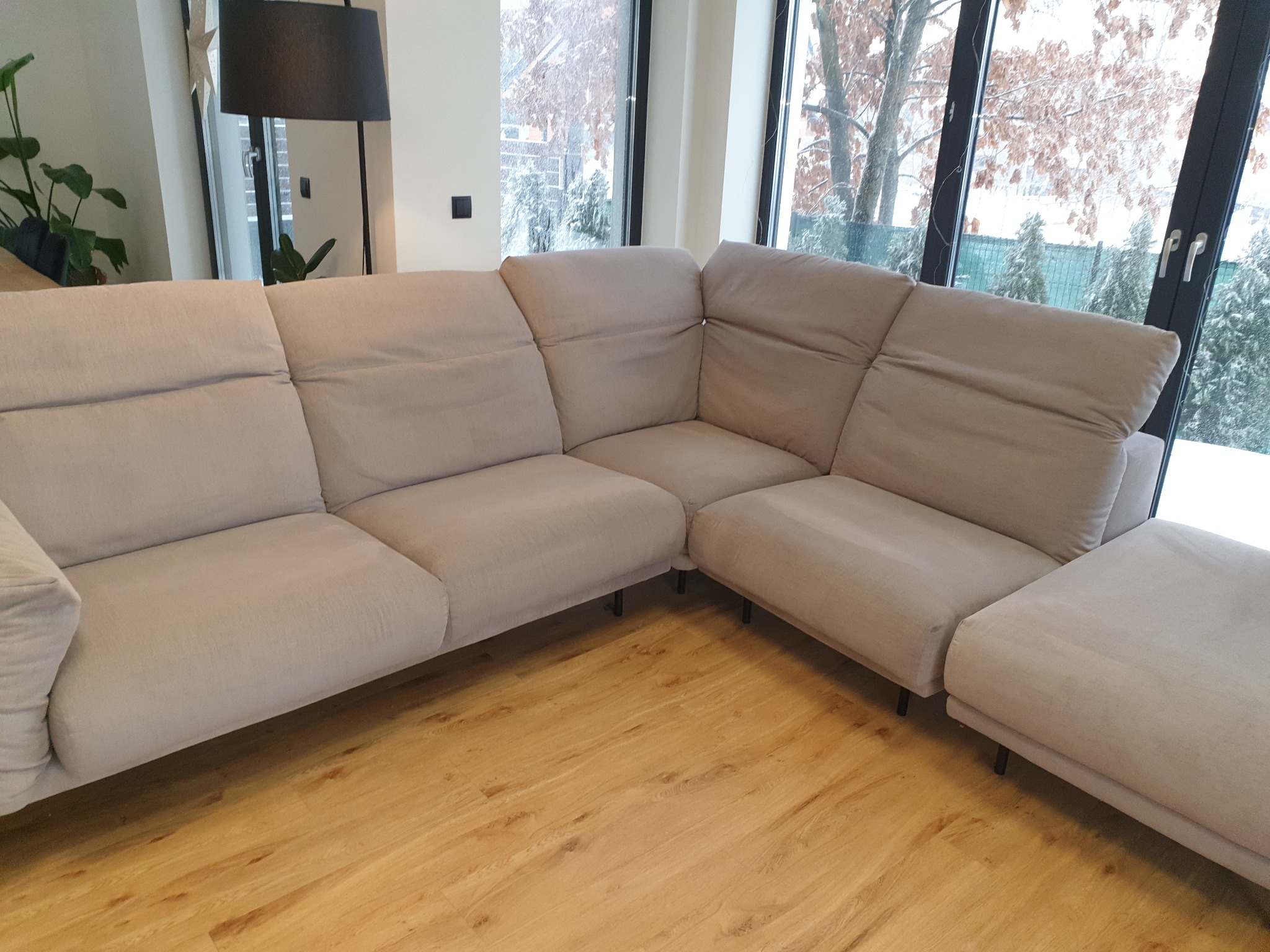 Jasny, narożny zestaw wypoczynkowy z zagłówkami, ustawiony na drewnianej podłodze w nowoczesnym wnętrzu z dużym oknem, za którym widać zimowy krajobraz.