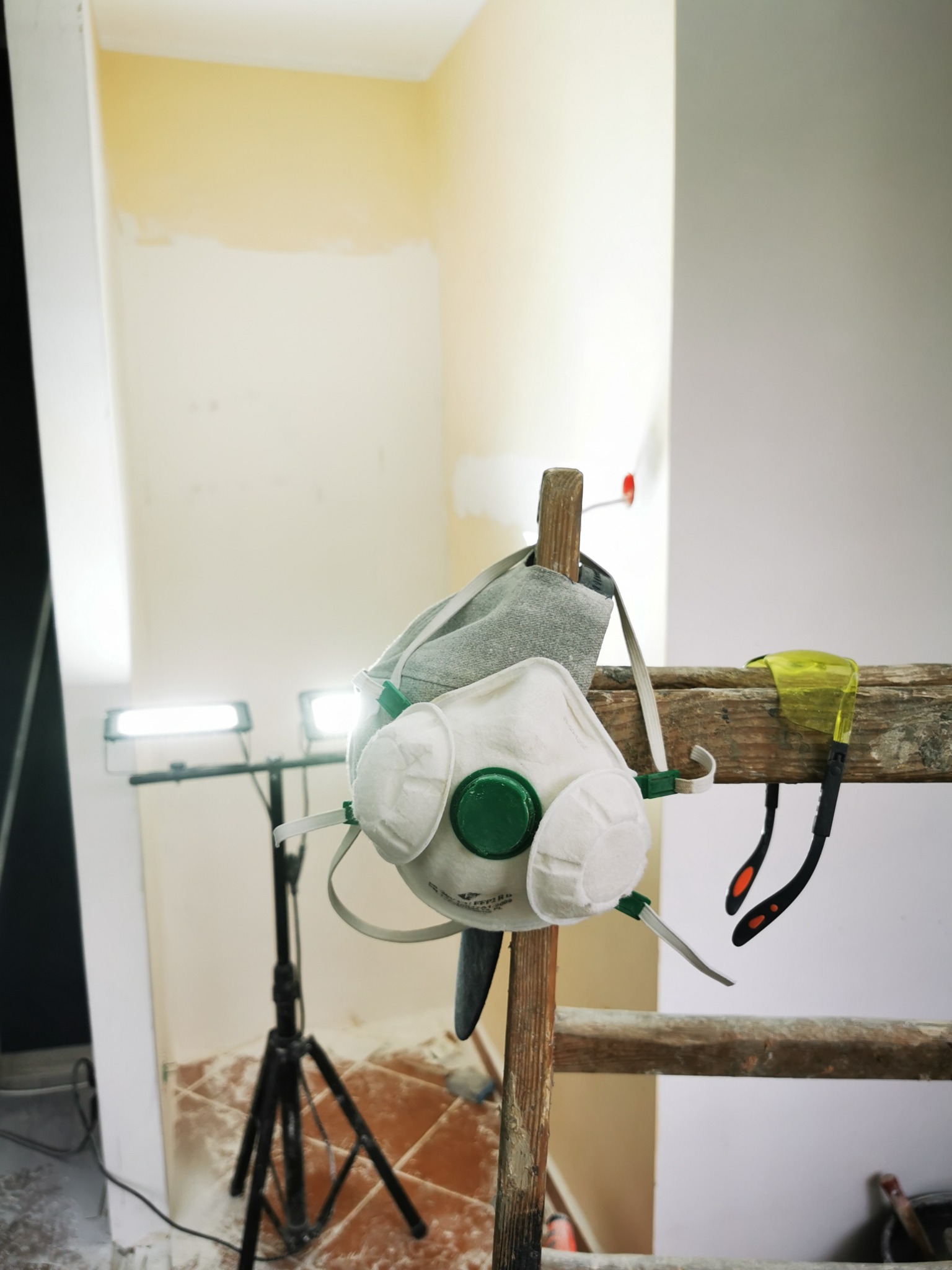 Maska ochronna i okulary wiszące na drewnianej drabinie w trakcie prac malarskich, widoczne niedomalowane ściany i lampa oświetleniowa.