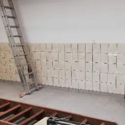 Kładzenie beżowych płytek kwadratowych na ścianie do połowy wysokości pomieszczenia, widoczna aluminiowa drabina i drewniana konstrukcja podłogowa.