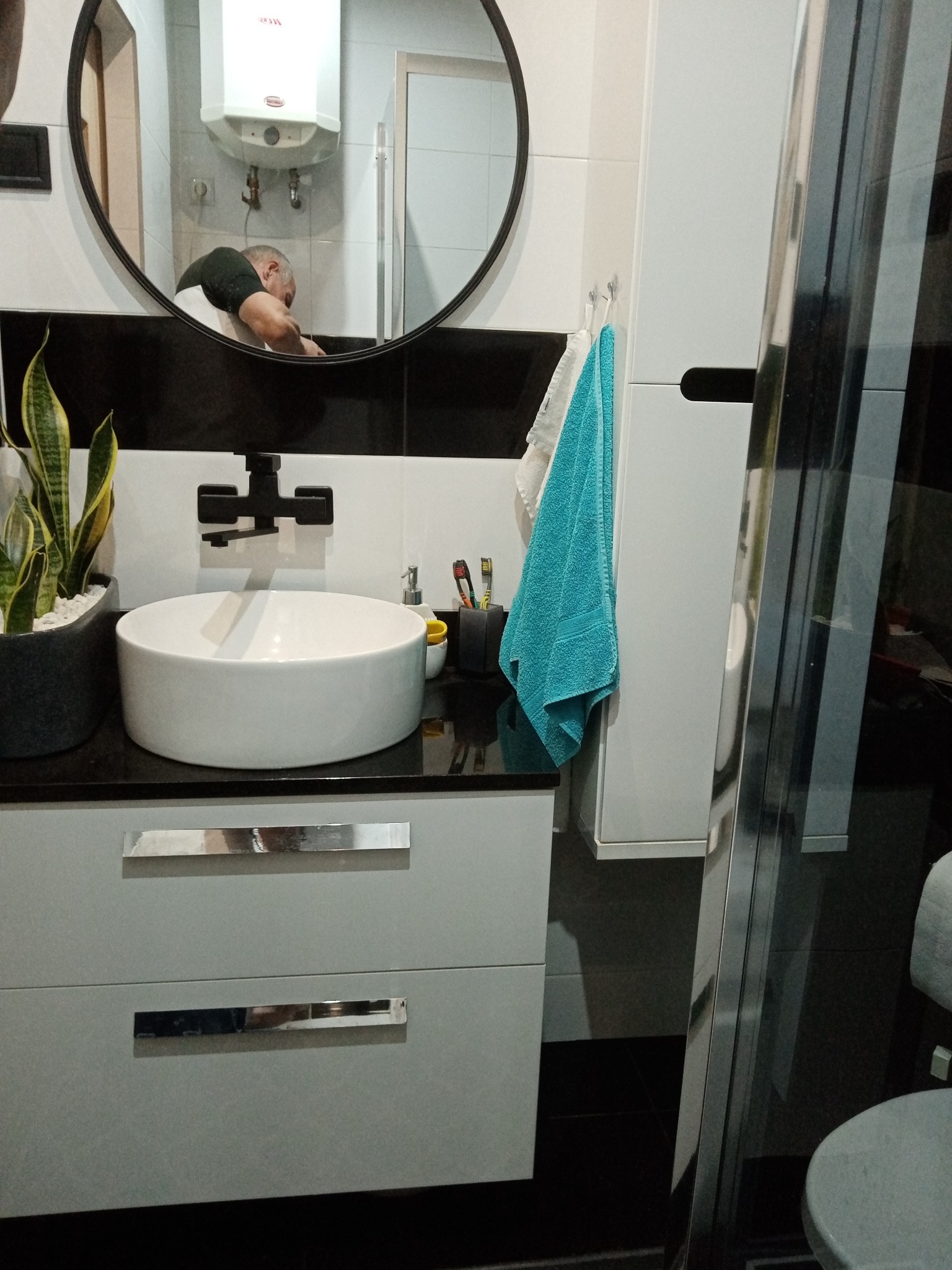 Nowoczesna łazienka z okrągłą umywalką na szafce z szufladami, czarną armaturą i lustrem, w którym odbija się osoba pracująca przy instalacji bojlera elektrycznego.