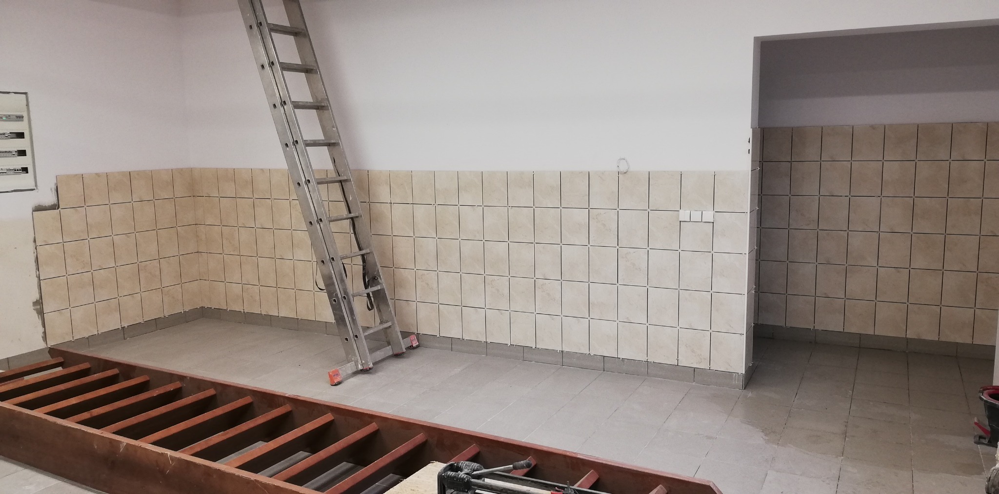 Kładzenie beżowych płytek kwadratowych na ścianie do połowy wysokości pomieszczenia, widoczna aluminiowa drabina i drewniana konstrukcja podłogowa.