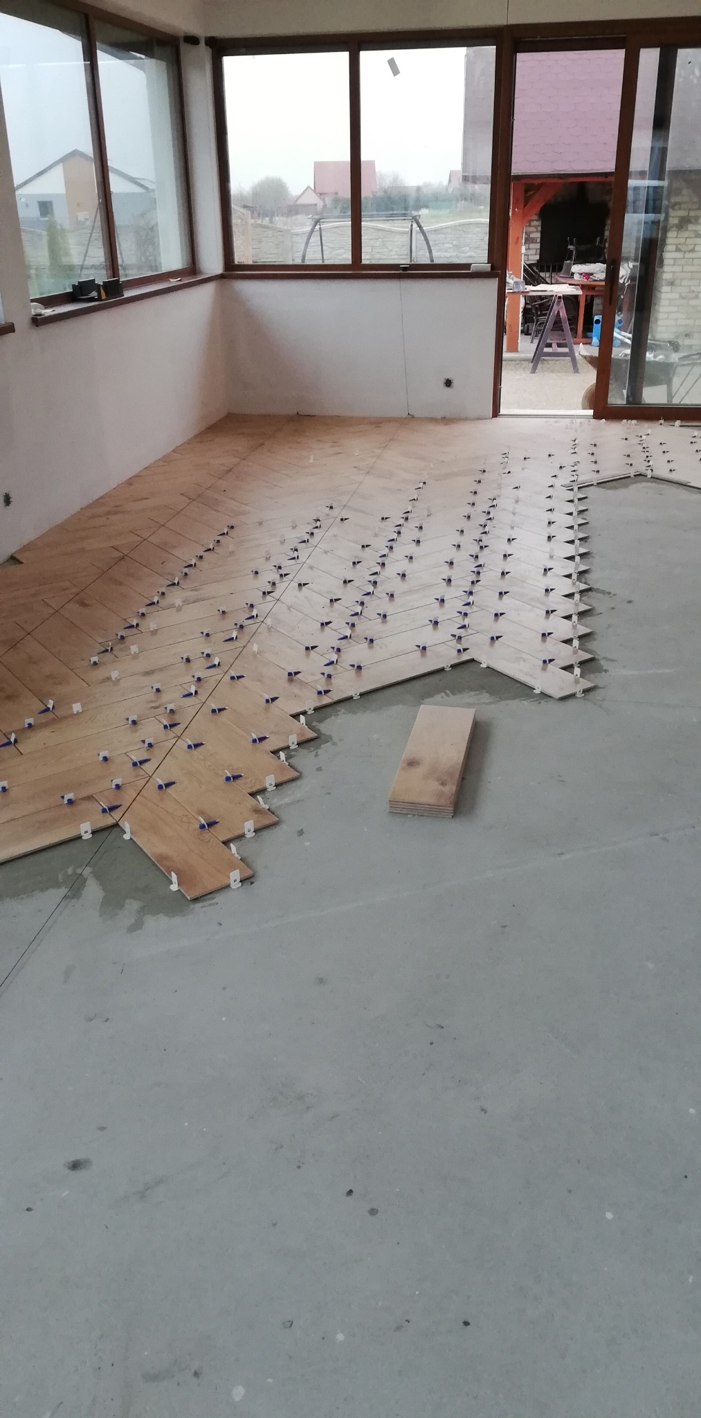 Układanie drewnopodobnych płytek podłogowych w jodełkę, z użyciem plastikowych krzyżyków dystansowych, na szarym, surowym podłożu w jasnym pomieszczeniu z dużymi oknami.