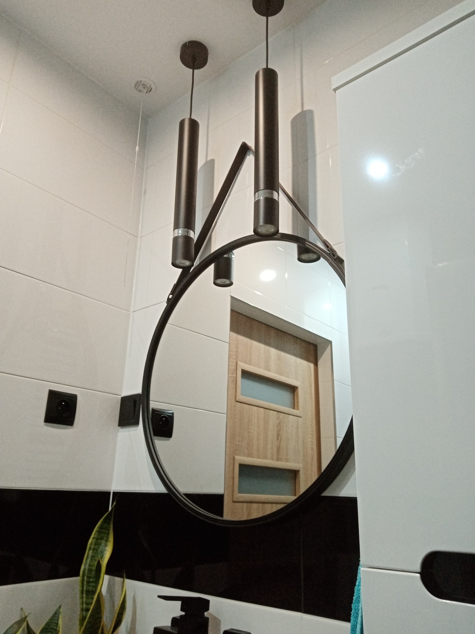 Nowoczesna łazienka z okrągłym lustrem wiszącym na metalowej ramie, oświetlonym trzema cylindrycznymi lampami; w tle widoczne drzwi z przeszkleniami.