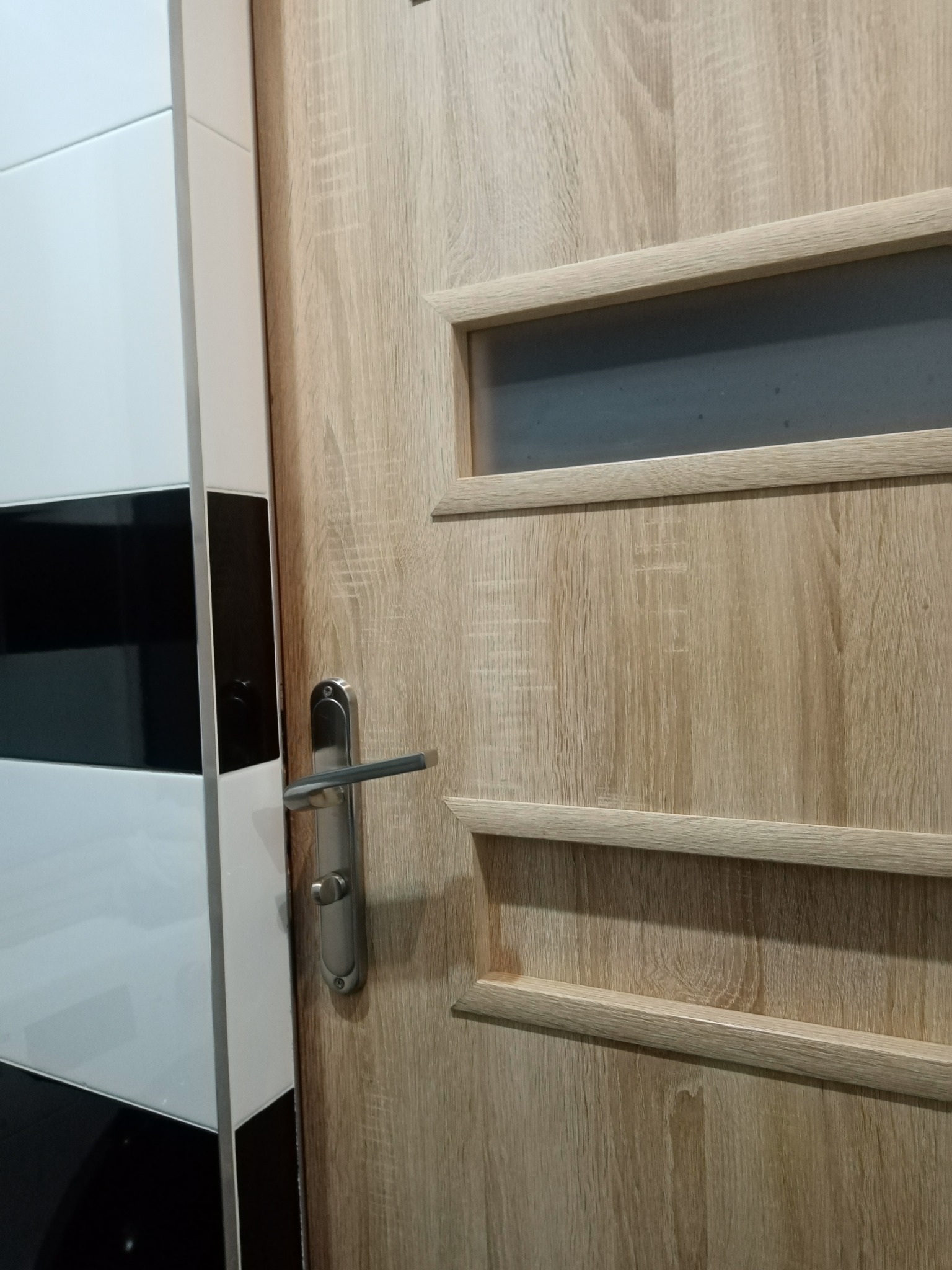 Zamknięte drzwi wewnętrzne w odcieniach drewna z metalową klamką i przeszklonym panelem, widoczne fragmenty czarno-białej ściany w tle.