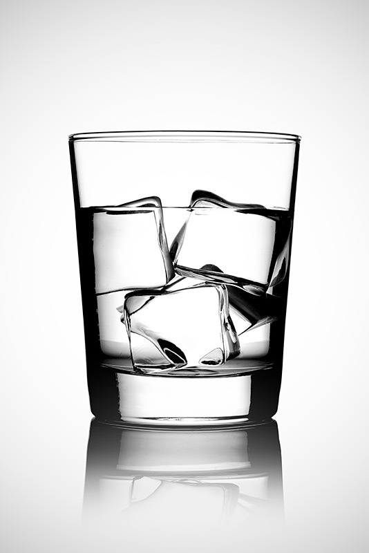Szklanka z wodą i kostkami lodu na białym tle z odbiciem, oświetlenie studyjne uwydatnia załamania światła w lodzie i wodzie.