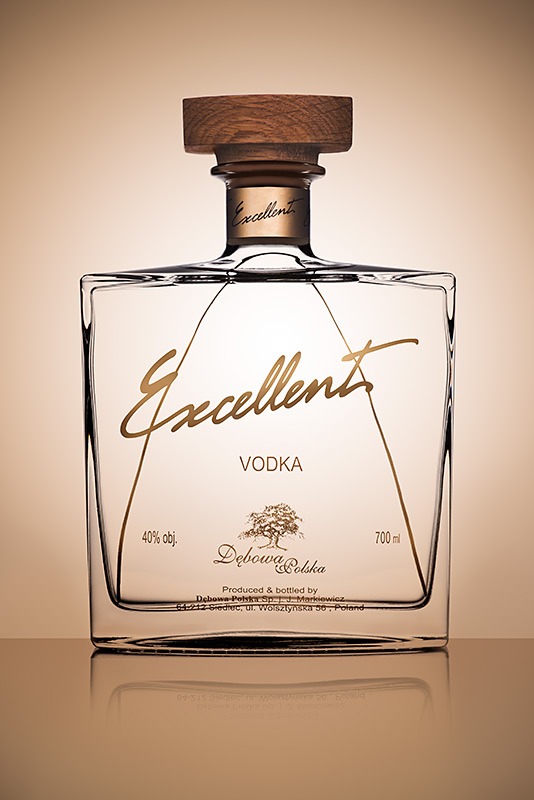 Kwadratowa butelka wódki Excellent Dębowa Polska z drewnianym korkiem, złote napisy i logo drzewa na etykiecie, odbicie na błyszczącym tle.
