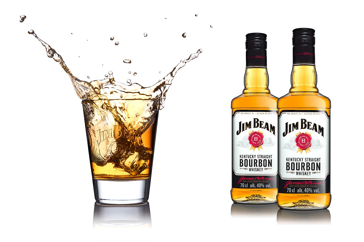 Kieliszek z kostkami lodu i tryskającym bourbonem Jim Beam obok dwóch butelek tego samego trunku na białym tle.
