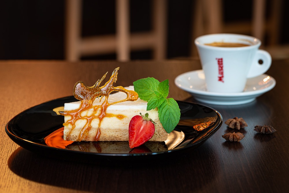 Kawałek sernika nowojorskiego na czarnym talerzu, udekorowany karmelem, truskawką i miętą, obok filiżanka espresso i anyż gwiazdkowy na ciemnym drewnianym stole.