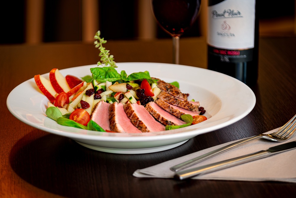 Elegancko podana sałatka z grillowaną kaczką, jabłkiem, pomidorami i sosem balsamicznym na białym talerzu, w tle kieliszek czerwonego wina i butelka Pinot Noir.