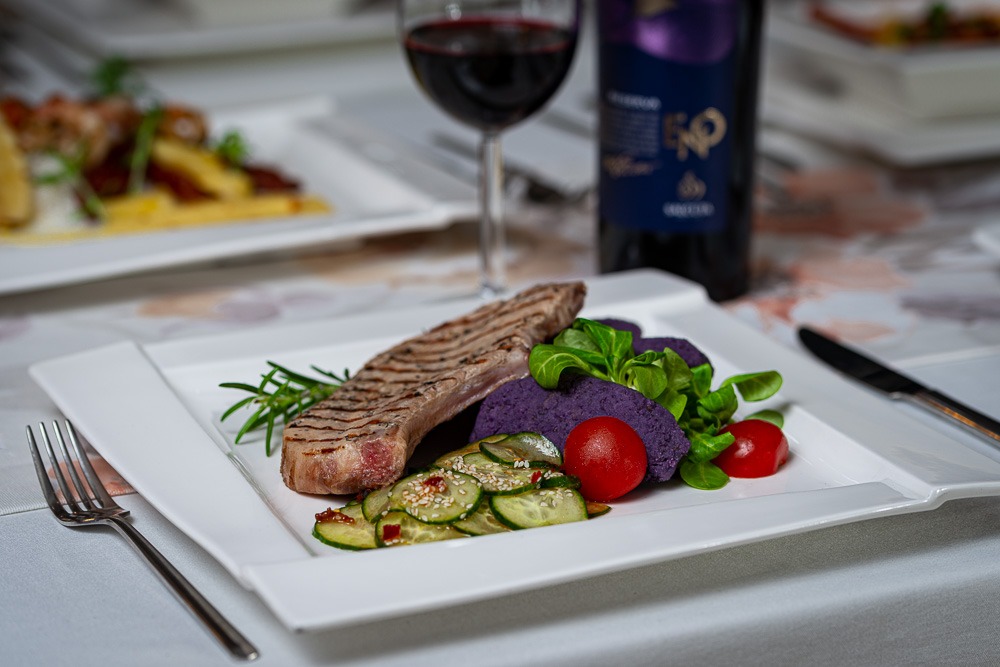 Elegancko podany stek z fioletowym puree ziemniaczanym, sałatką z ogórków i pomidorkami cherry na białym kwadratowym talerzu, w tle kieliszek czerwonego wina i butelka wina.