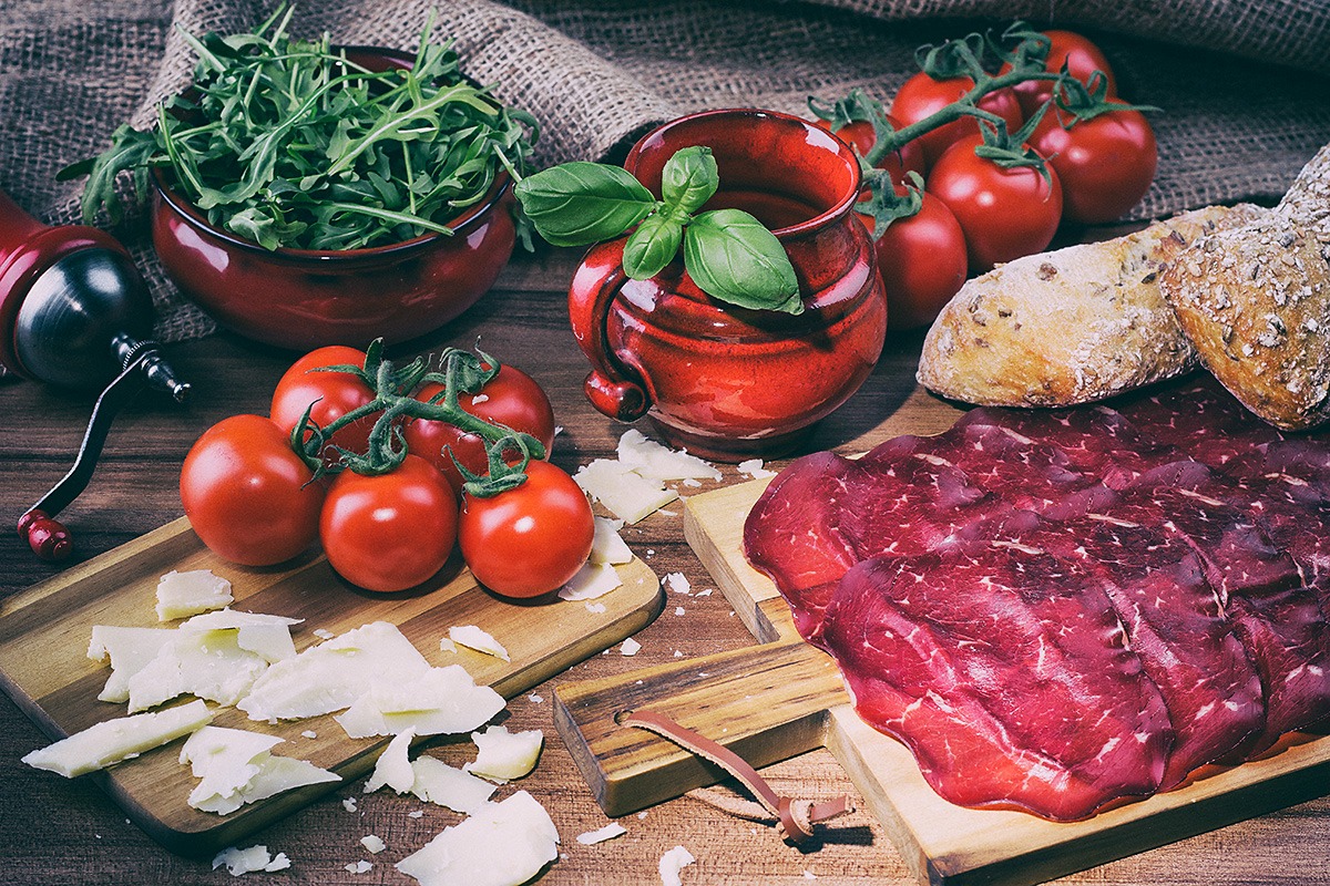 Kompozycja kulinarna: rukola w czerwonej misce, czerwony dzbanek z bazylią, pomidory na gałązce, ser w płatkach, pieczywo i plastry suszonej wołowiny na drewnianych deskach, z młynkiem do pieprzu...