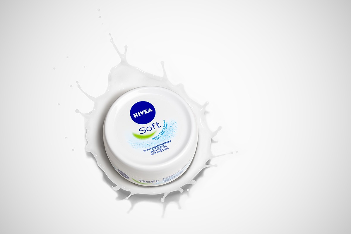 Pojemnik kremu Nivea Soft otoczony rozpryskującym się białym płynem na jasnym tle, prezentacja produktu z perspektywy z góry.