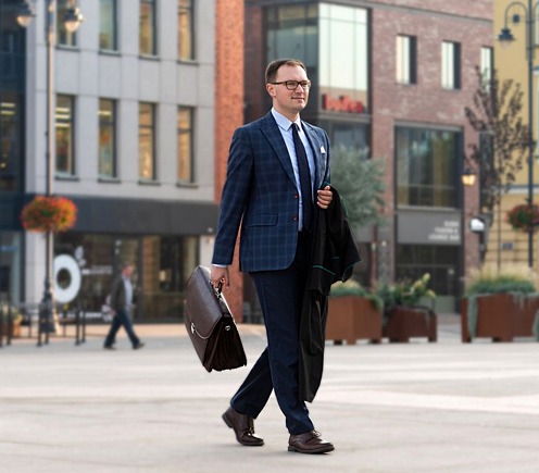 Mężczyzna w okularach, granatowym garniturze w kratę, z teczką i togą adwokacką w ręku, idący po placu miejskim.