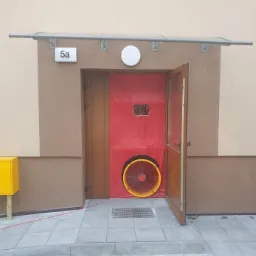 Wejście do budynku z zamontowanym wentylatorem w czerwonej płachcie, test szczelności drzwi, widoczny numer budynku 5a i żółta skrzynka gazowa