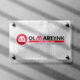 Szyld firmy 'OLMARTYNK TYNKI AGREGATEM' na przezroczystej płycie, przymocowanej do szarej ściany, oświetlonej promieniami słonecznymi.