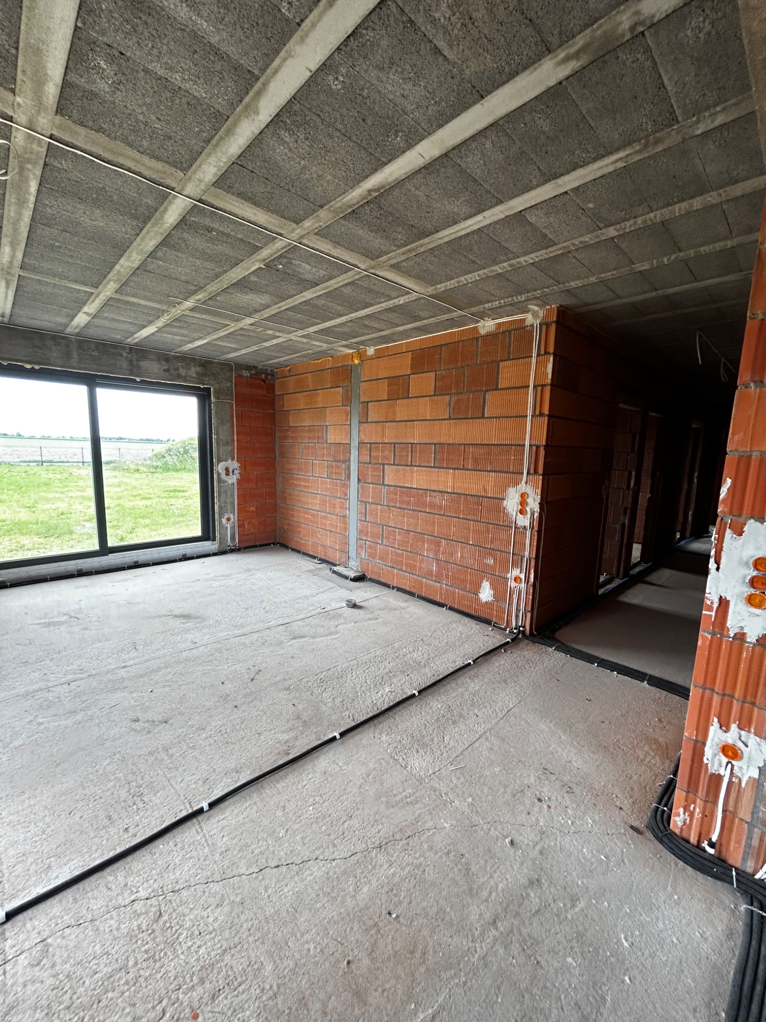 Surowy stan wnętrza budynku w trakcie budowy z widocznymi przewodami elektrycznymi ułożonymi na podłodze i wyprowadzonymi punktami instalacji w ścianach z cegły ceramicznej.
