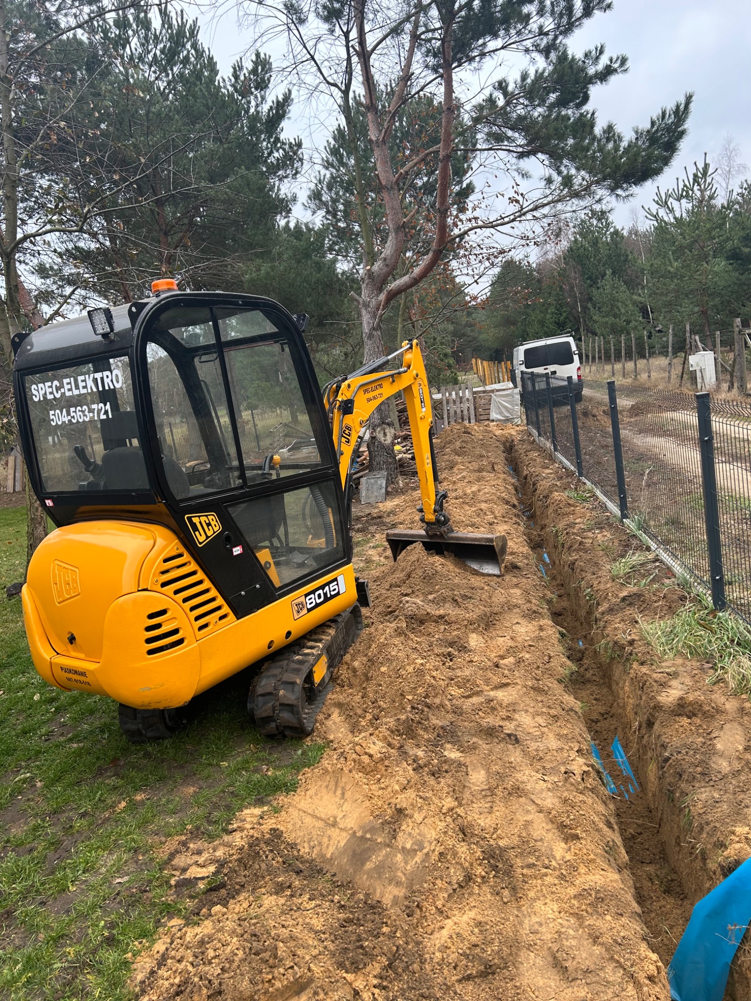 Żółta mini koparka JCB 8015 obok wykopanego rowu z niebieskimi rurami w ziemi, widoczny numer telefonu na koparce i samochód dostawczy w tle.