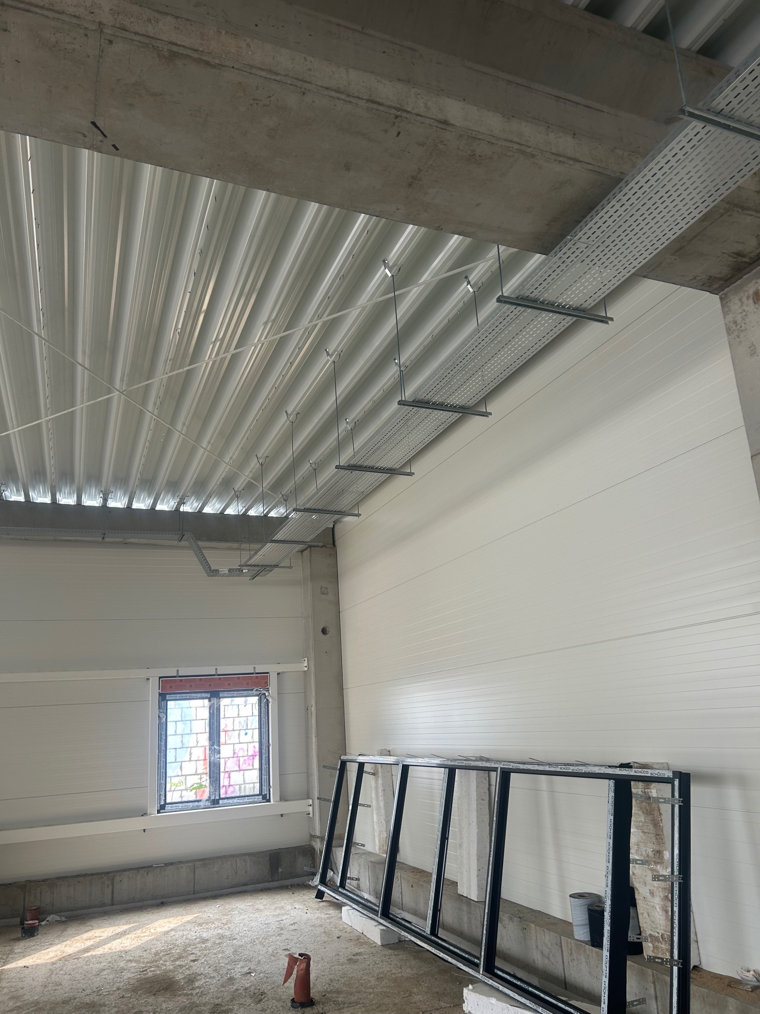Montaż korytek kablowych pod sufitem w nowym budynku. Widoczne elementy konstrukcyjne i okno. Szary beton, białe ściany i sufit z blachy trapezowej.