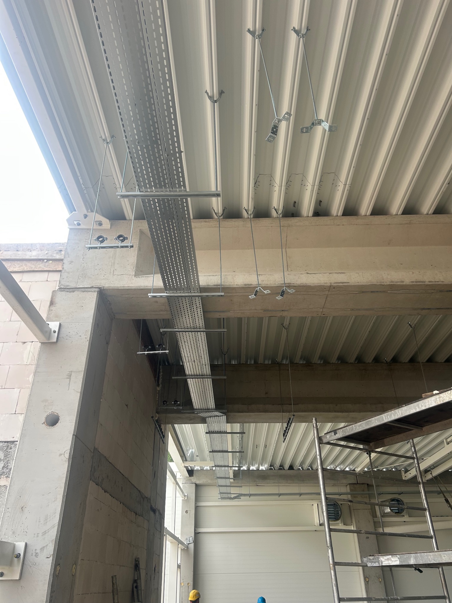 Montaż perforowanych koryt kablowych pod sufitem hali przemysłowej, widoczne elementy mocujące i wsporniki. Prace instalacyjne w surowym stanie budynku.