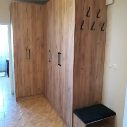 Komplet szaf narożnych z drewnopodobnym wykończeniem, czarnymi uchwytami i wieszakami na ścianie, uzupełniony o tapicerowane siedzisko z półkami w przedpokoju.