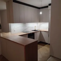 Nowoczesna kuchnia w odcieniach bieli i drewna z blatem imitującym drewno i marmurowym panelem nad blatem, złota bateria przy czarnym zlewie, podłoga ułożona w jodełkę.