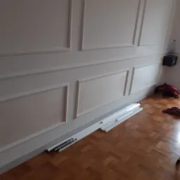 Pokój z panelami ściennymi w trakcie montażu, widoczne listwy przypodłogowe na podłodze, drabina, butelka wody i torba w tle.