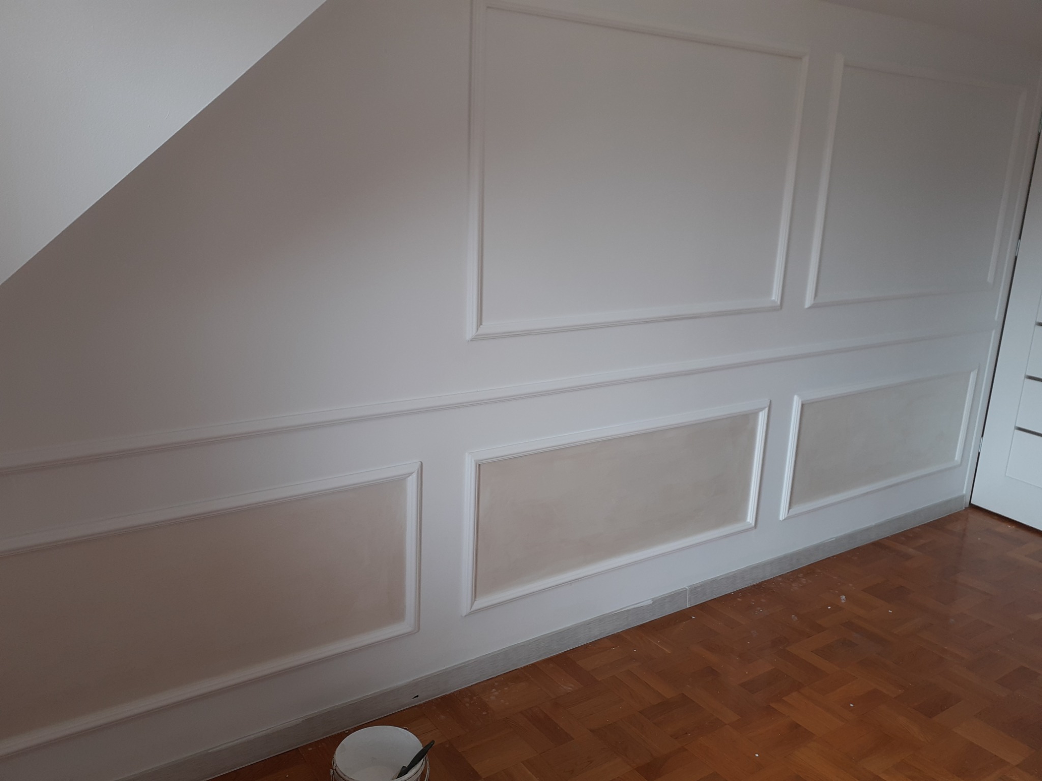 Ściana w pokoju po częściowym wykończeniu z ozdobnymi panelami, górne panele pomalowane na biało, dolne w odcieniach beżu, widoczny fragment poddasza, parkiet na podłodze i wiaderko z farbą w rogu.