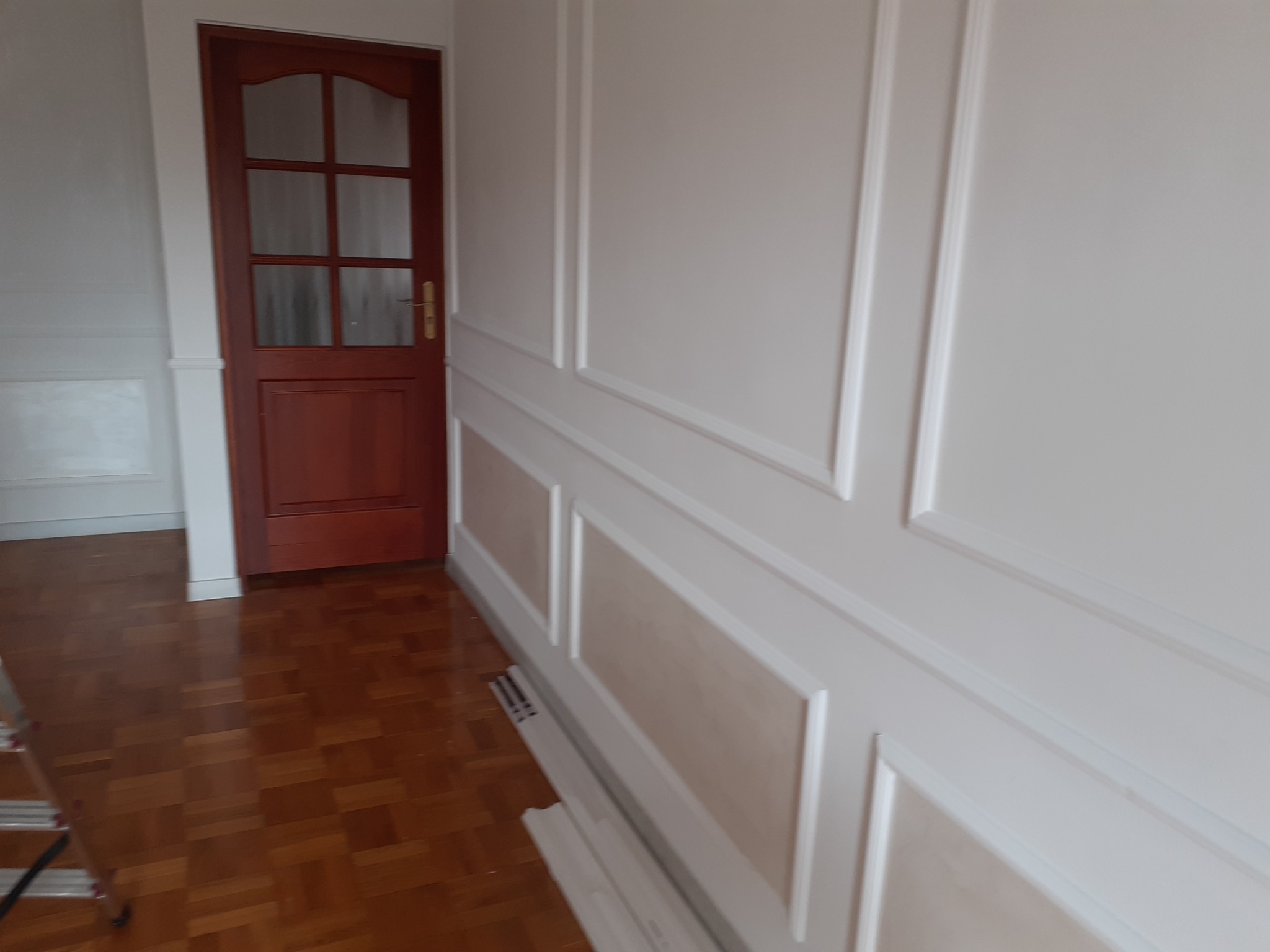 Świeżo pomalowane, białe ściany z dekoracyjnymi listwami i panelami, brązowe drzwi z szybami, parkiet na podłodze, widoczne listwy przypodłogowe i fragment drabiny