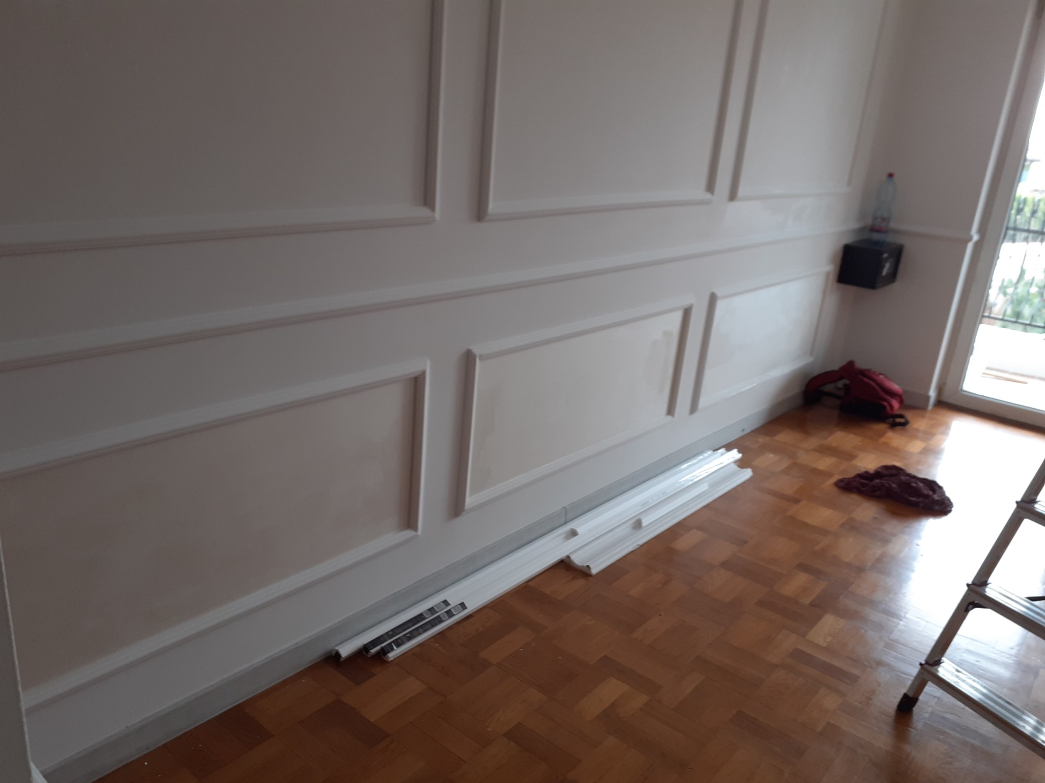 Pokój z panelami ściennymi w trakcie montażu, widoczne listwy przypodłogowe na podłodze, drabina, butelka wody i torba w tle.