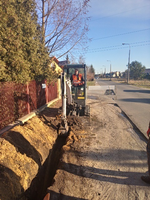 Mini koparka wykopuje wąski rów wzdłuż ogrodzenia, operator w kamizelce odblaskowej, widok na ulicę i zabudowania w tle.