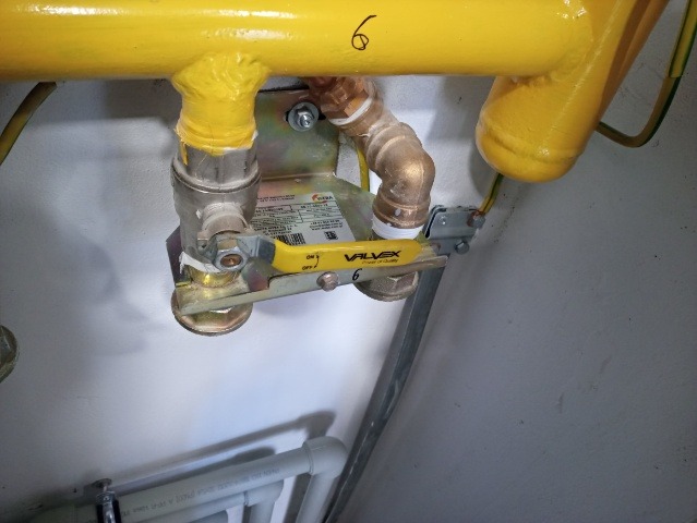 Żółta rura gazowa z zaworem VALVEX i numerem '6', widoczne połączenia i elementy instalacji, fragment grzejnika PCV w tle.