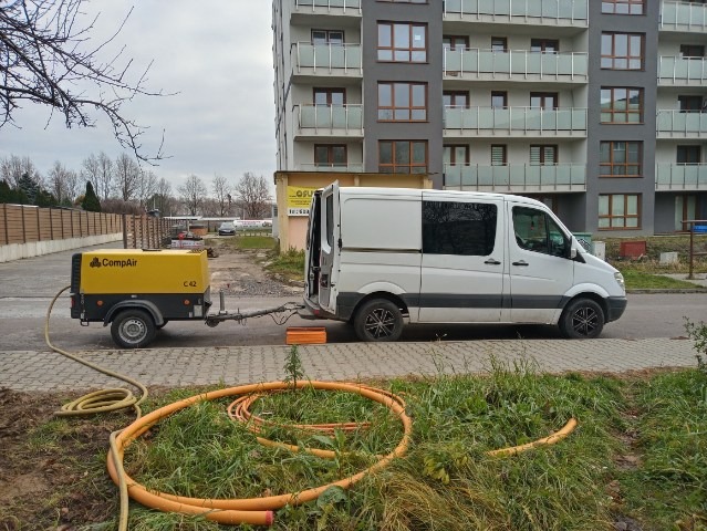 Żółty kompresor powietrza CompAir C42 na przyczepce podłączony do białego furgonu na tle bloku mieszkalnego, z pomarańczowymi rurami leżącymi na ziemi.