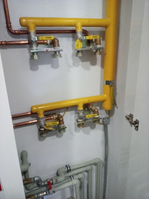 Żółte i miedziane rury instalacji gazowej z zaworami, zamontowane na białej ścianie, widoczne elementy instalacji wodnej w dolnej części kadru.