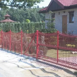 Czerwone ogrodzenie panelowe z bramą przesuwną na tle domu z białej cegły i zieleni. Widoczny fragment chodnika i trawnika wewnątrz posesji.