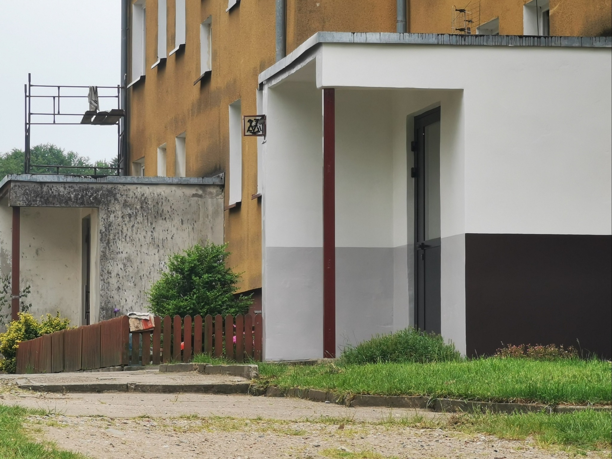 Odnowiona elewacja wejścia do budynku, z białymi ścianami i szarym pasem na dole, brązowe filary oraz ciemne drzwi wejściowe, widoczny fragment starej, żółtej elewacji z symbolem na ścianie, mały...