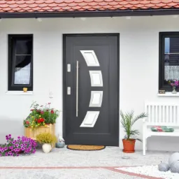 Nowoczesne, grafitowe drzwi zewnętrzne z czterema białymi, ukośnymi panelami, długim, srebrnym uchwytem i dwoma małymi oknami po bokach. Przed drzwiami kwiaty i biała ławka.